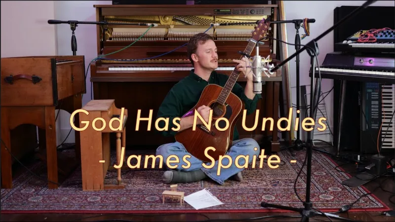 James Spaite sitting cross-legged on a patterned rug with an acoustic guitar, an upright piano and microphones behind him.