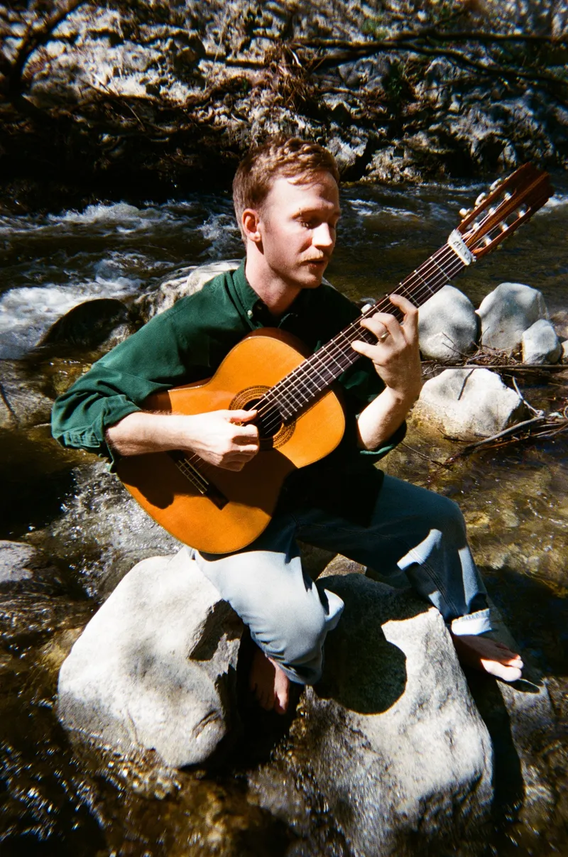 James Spaite seated on a rock in a creek, eyes on the fretboard, both hands on a classical guitar.